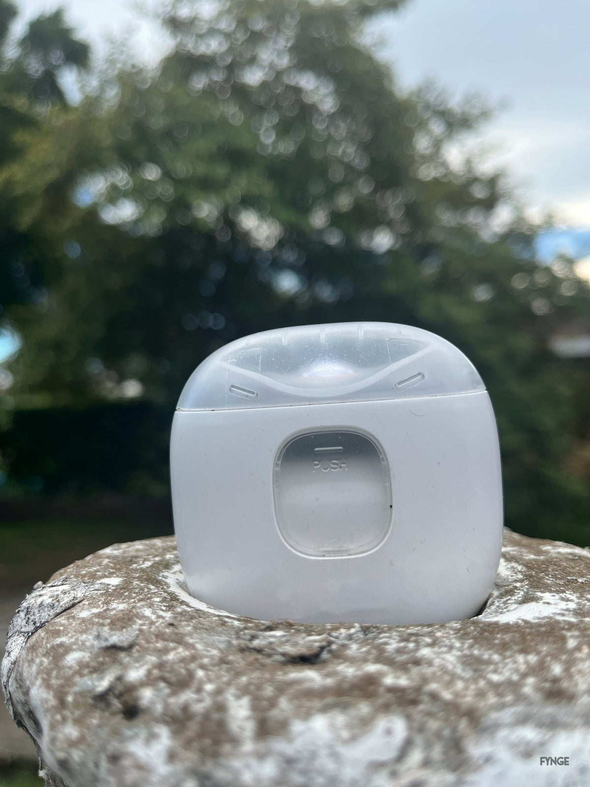 Back of the FYNGE trimmer on a stone post, showing the PUSH clippings door, trees behind